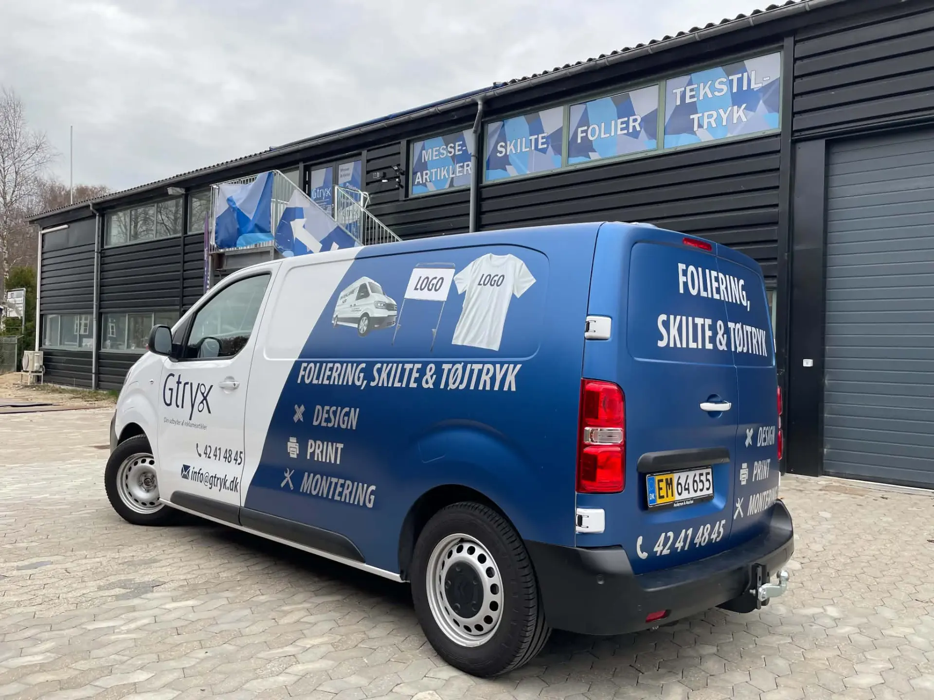 Gtryks egen blå og hvide firmabil med foliering, parkeret foran virksomhedens bygning i Kvistgård med skilte for messeartikler, skilte, folier og tekstiltryk.
