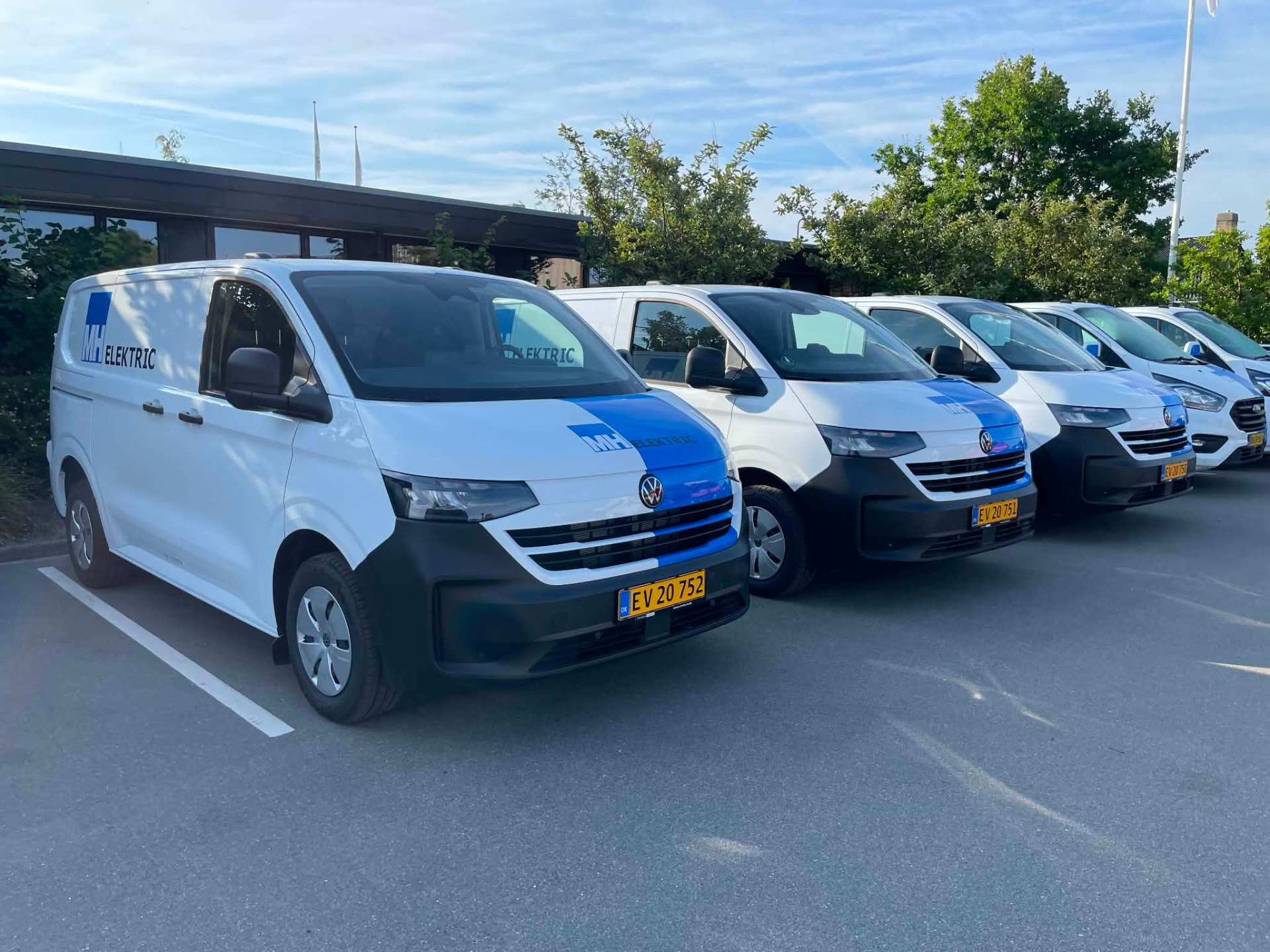 Flåde af hvide varevogne med blå MH Elektric-logofolie på sider og motorhjelm, parkeret side om side.