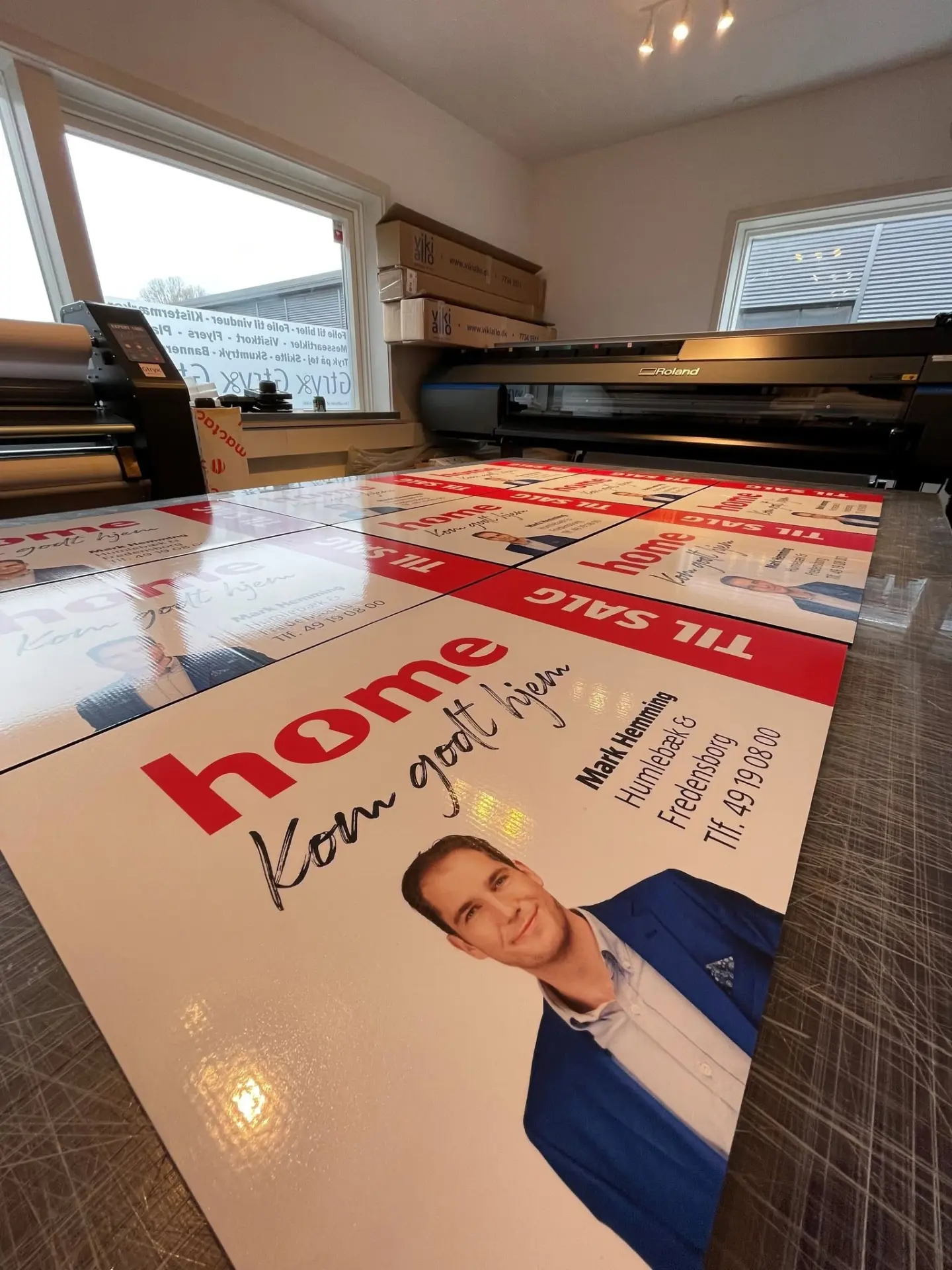 Indstik fra et trykkeri, hvor store “Til salg”-plakater med mæglerlogo og kontaktoplysninger ligger klar på produktionsoverfladen foran en storformatprinter.